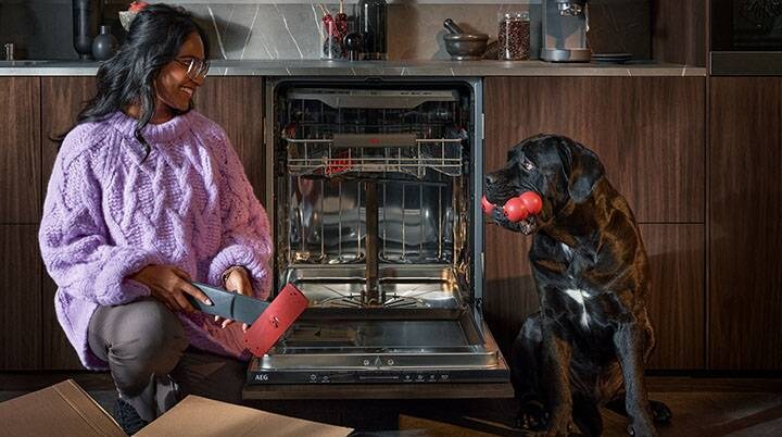 Žena vybaluje náhradní díly do myčky a usmívá se na černého psa s hračkou v tlamě